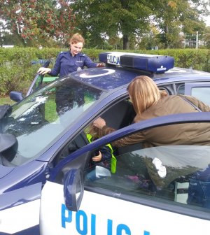 Dziewczynka siedzi na fotelu kierowcy w policyjnym radiowozie, obok przy samochodzie stoi jej mama. Natomiast od strony pasażera przy drzwiach stoi policjantka.