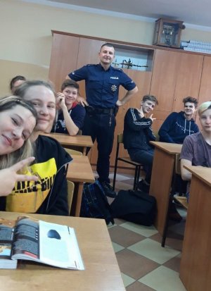 Dzielnicowy mł. asp. Michał Starczewski stoi na środku klasy, a wokół niego stoliki szkolne, w których siedzą uczniowie.