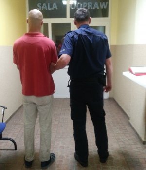 Zatrzymany 31-latek z lewej strony, z prawej policjant. Mężczyźni stoją na korytarzu w budynku Komendy Powiatowej Policji w Żninie. Policjant trzyma zatrzymanego pod rękę