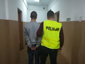 Korytarz w Komendzie Powiatowej Policji w Żninie. Z prawej strony policjant, z lewej zatrzymany mężczyzna z kajdankami na rękach.