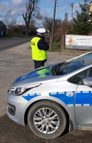 Policjant ruchu drogowego wykonuje pomiar prędkość przy użyciu laserowego miernika. Z prawej strony zaparkowany policyjny radiowóz.